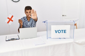 Young handsome hispanic man at political campaign election holding england flag with open hand...
