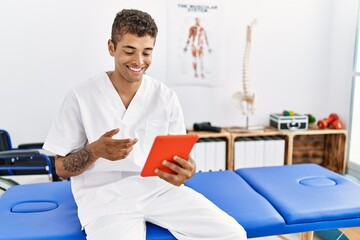 Young hispanic man working as physiotherapist doing videocall at physiotherapy room