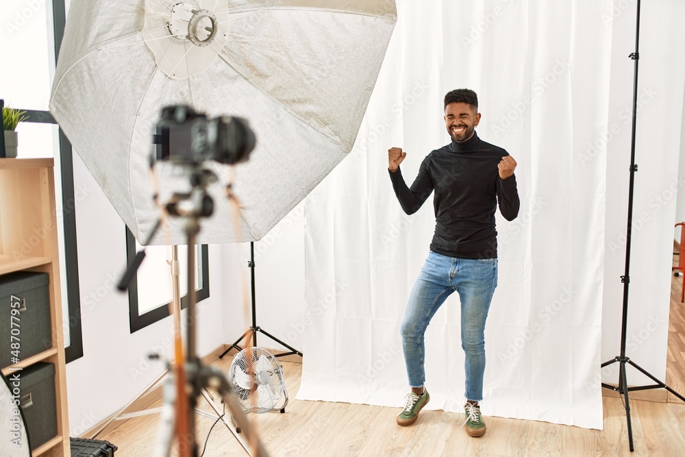 Poster young hispanic man with beard posing as model at photography studio very happy and excited doing win