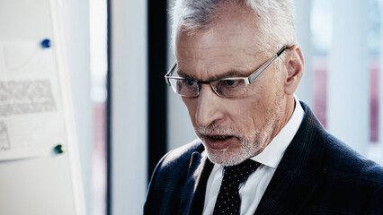 Naklejka na ściany i meble Middle aged businessman in eyeglasses standing near blurred flip chart in office