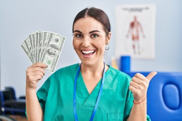 Young hispanic woman holding dollars banknotes working at pain recovery clinic pointing thumb up to the side smiling happy with open mouth