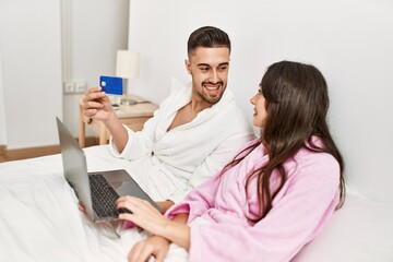 Young hispanic couple using laptop and credita card lying in bed at home.