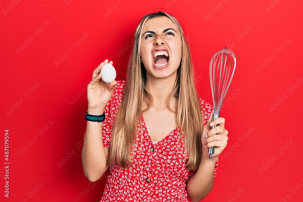 Sticker beautiful hispanic woman holding egg and baker whisk angry and mad screaming frustrated and furious,