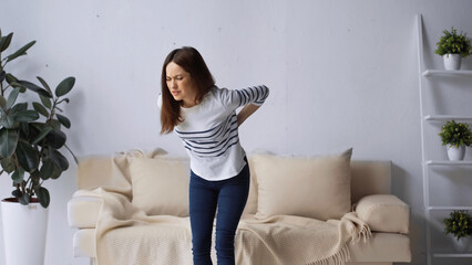 brunette woman with closed eyes standing near sofa and touching painful back - Powered by Adobe