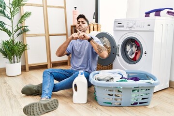 Young hispanic man putting dirty laundry into washing machine rejection expression crossing fingers doing negative sign