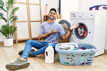 Young hispanic man putting dirty laundry into washing machine inviting to enter smiling natural with open hand