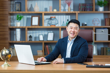 Successful asian businessman looking at camera and smiling, man working in classic office at desk, with laptop, looking at camera portrait - obrazy, fototapety, plakaty
