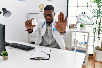 Young african american doctor man wearing doctor uniform holding prescription pills at the clinic with open hand doing stop sign with serious and confident expression, defense gesture