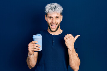 Young hispanic man with modern dyed hair drinking a take away cup of coffee pointing thumb up to the side smiling happy with open mouth