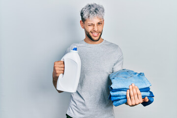 Young hispanic man with modern dyed hair holding jeans for laundry and detergent bottle winking...