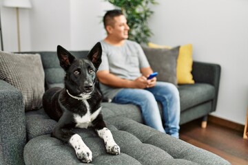 Young hispanic man using smartphone sitting on the sofa with dog at home.