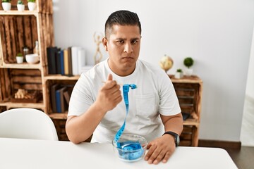 Young hispanic man unhappy eating tape measure as a diet concept at home.