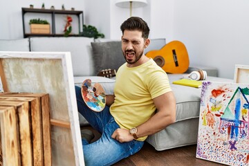 Young man with beard painting canvas at home winking looking at the camera with sexy expression,...