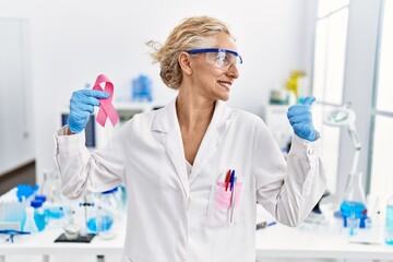 Middle age blonde woman working at laboratory looking for breast cancer cure pointing thumb up to...