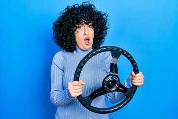 Young middle east woman holding steering wheel in shock face, looking skeptical and sarcastic,...