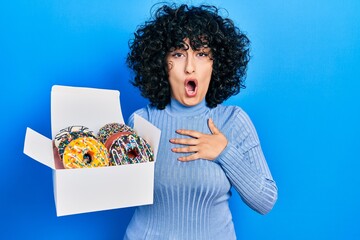 Young middle east woman holding tasty colorful doughnuts box scared and amazed with open mouth for...