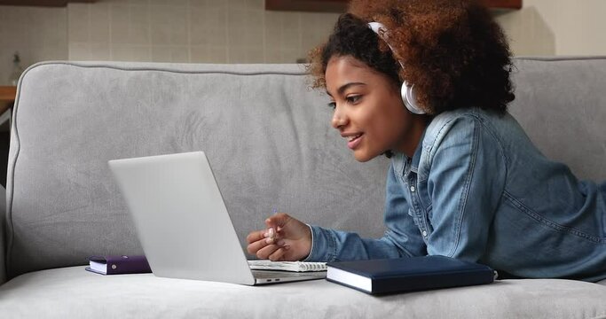 African teenage girl lying on couch studying from home use computer and video call application, take notes, do college assignment, prepare for university admission or exams. E-learning, tech concept