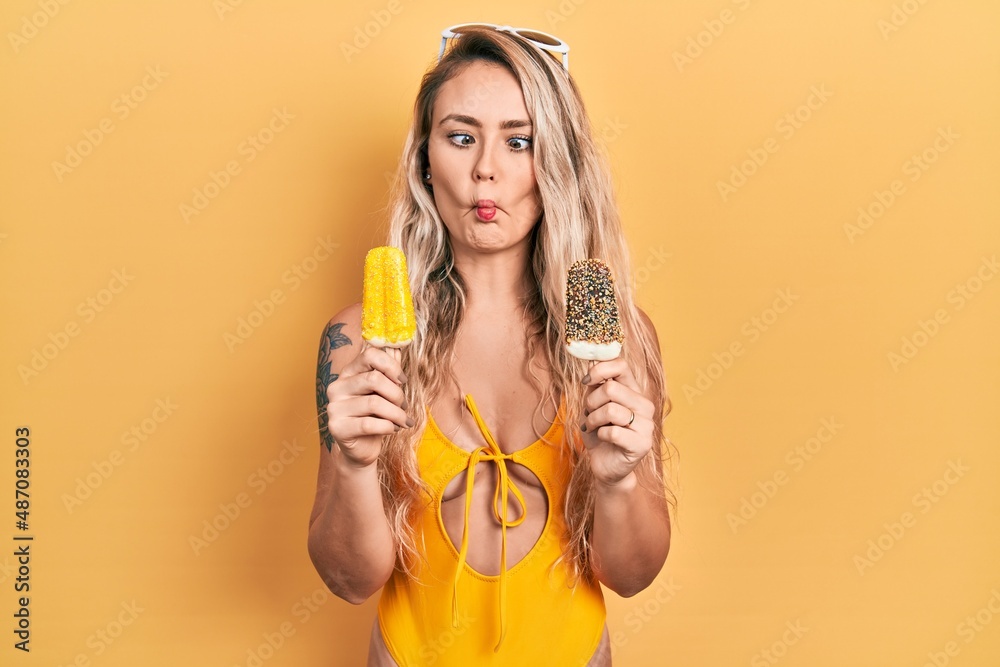 Poster Beautiful young blonde woman wearing summer swimsuit holding ice creams making fish face with mouth and squinting eyes, crazy and comical.