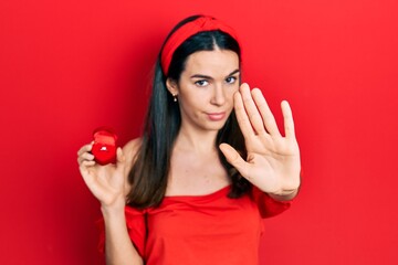 Young brunette woman holding engagement ring for proposal with open hand doing stop sign with...