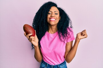 Middle age african american woman holding mango screaming proud, celebrating victory and success...