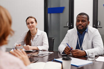 Two young intercultural healthcare experts in lab coats consulting colleagues about frequently...