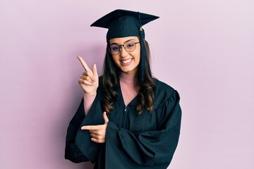 Young hispanic woman wearing graduation cap and ceremony robe smiling and looking at the camera pointing with two hands and fingers to the side.