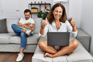 Hispanic middle age couple at home, woman using laptop success sign doing positive gesture with hand, thumbs up smiling and happy. cheerful expression and winner gesture.