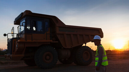 Silhouette of a truck driver wearing a helmet and safety vest with a large truck in the background.
