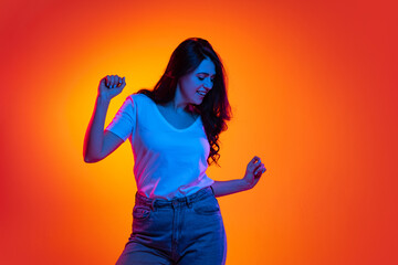 Excited young beautiful caucasian girl with long dark hair dancing isolated on orange background in neon light, filter. Concept of emotions, ads, sale