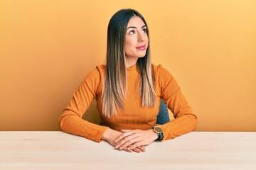 Young hispanic woman wearing casual clothes sitting on the table smiling looking to the side and staring away thinking.