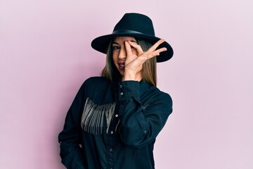 Young hispanic woman wearing cowgirl style smiling happy doing ok sign with hand on eye looking through fingers