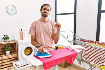 Young hispanic man ironing clothes at home pointing up looking sad and upset, indicating direction...