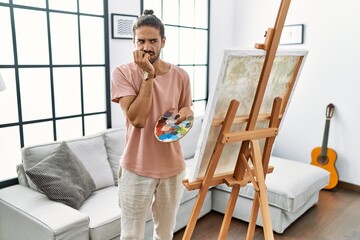 Young hispanic man with beard painting on canvas at home looking stressed and nervous with hands on mouth biting nails. anxiety problem.