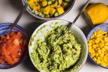 Bowls in a kitchen filled with guacamole, corn, tomatoes, mango and cucumber.
