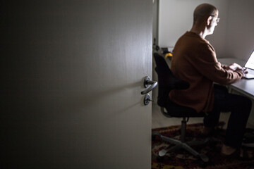 A man sits at a computer in a home office in the dark.