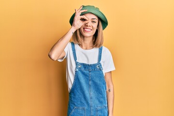 Young caucasian blonde woman wearing denim jumpsuit and hat with 90s style doing ok gesture with hand smiling, eye looking through fingers with happy face.