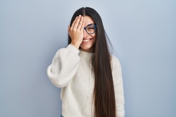 Young hispanic woman wearing casual sweater over blue background covering one eye with hand, confident smile on face and surprise emotion.