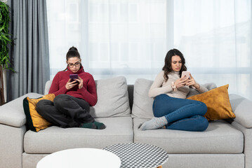 Two young woman roommates sitting on the sofa at home mad after argue not talking. Females addiction to internet and online messaging, surfing the net on their smartphones.