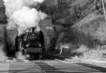 Steam train with historic locomotive and coming to light out of “Schloßberg Tunnel“ at railroad crossing in Arnsberg Sauerland Germany on Ruhr Valley line. Vintage black and historic railway scene.