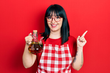 Young hispanic woman wearing cook apron holding olive oil can smiling happy pointing with hand and...