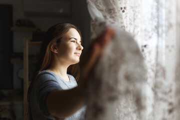 Fototapeta premium Pretty young woman in apartment opening window curtains after wake up, enjoying morning sunshine .Mental health concept .