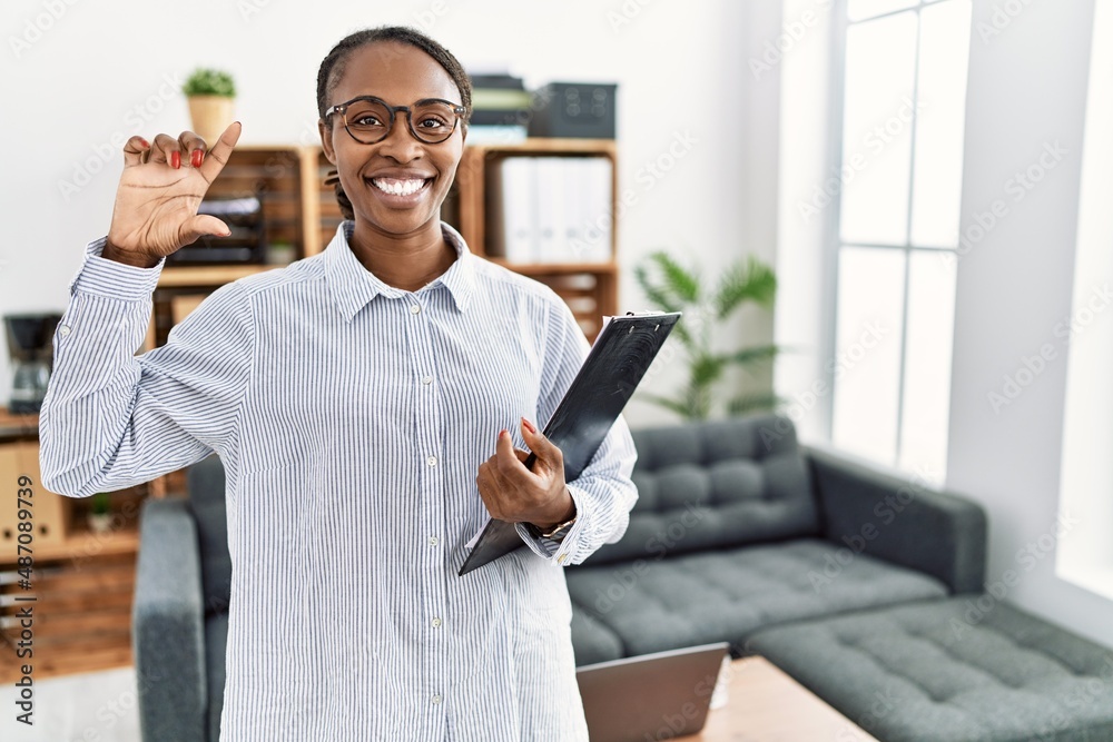 Canvas Prints African woman working at psychology clinic smiling and confident gesturing with hand doing small size sign with fingers looking and the camera. measure concept.