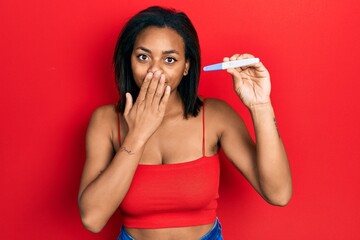 Naklejka premium Young african american girl holding pregnancy test result covering mouth with hand, shocked and afraid for mistake. surprised expression
