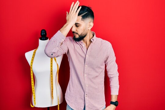 Handsome tailor man with beard standing by manikin surprised with hand on head for mistake, remember error. forgot, bad memory concept.