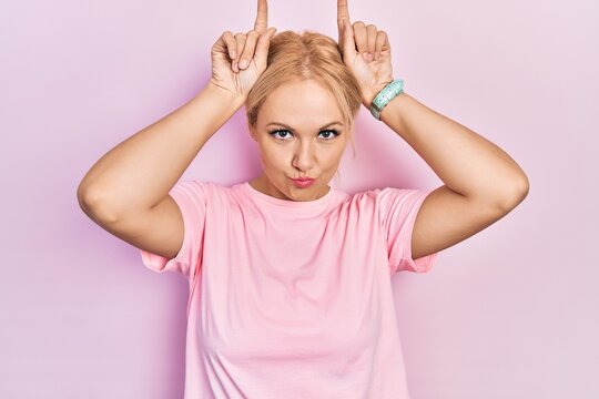 Young Blonde Woman Wearing Casual Pink T Shirt Doing Funny Gesture With Finger Over Head As Bull Horns