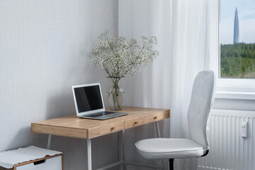 Work desk with a laptop on the table and a vase of flowers, an armchair and a view of a skyscraper in a field from the window