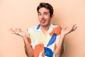 Young caucasian man isolated on beige background doubting and shrugging shoulders in questioning...
