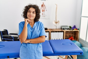 Young hispanic physiotherapist woman working at pain recovery clinic serious face thinking about question with hand on chin, thoughtful about confusing idea