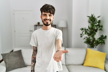 Hispanic man with beard at the living room at home smiling cheerful offering palm hand giving assistance and acceptance.