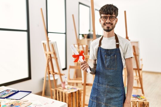 Young hispanic artist man smiling happy holding diploma at art studio.